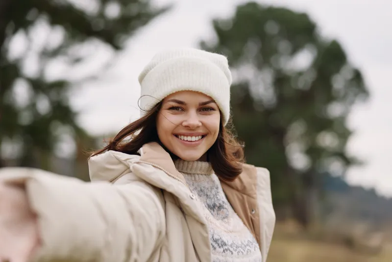 Smiling woman in winter outdoors, wearing a beanie and coat, reaching her arm toward the camera for a candid selfie against background