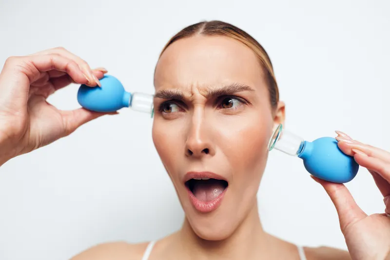 Young woman using blue suction cups on ears with confused expression on white background. Concept of alternative therapy, ear cleaning, and health care.