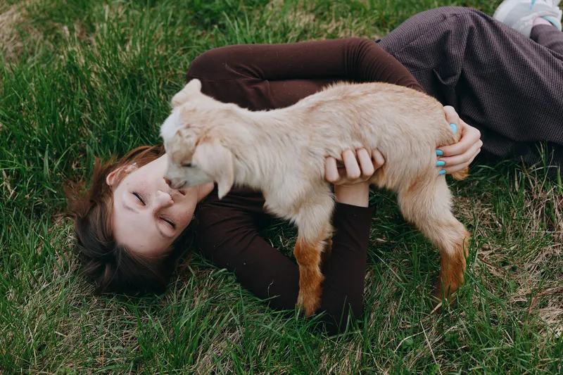 Joyful woman outdoors, lying on lush grass, embracing a playful goat kid, warm sunlight, relaxed bond, natural colors, tranquil countryside vibe