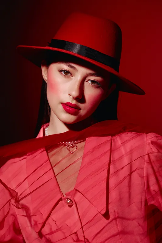 Woman in red hat and blouse poses under dramatic red lighting. Face makeup features bold lips and rosy cheeks in a creative beauty fashion style.