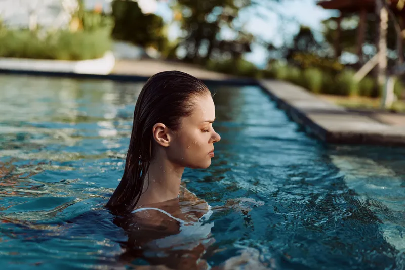 Relaxed woman glides in a sunlit pool, water ripples around her shoulders, presenting a serene lifestyle moment isolated by a quiet pool edge, signaling personal downtime and gentle outdoor leisure