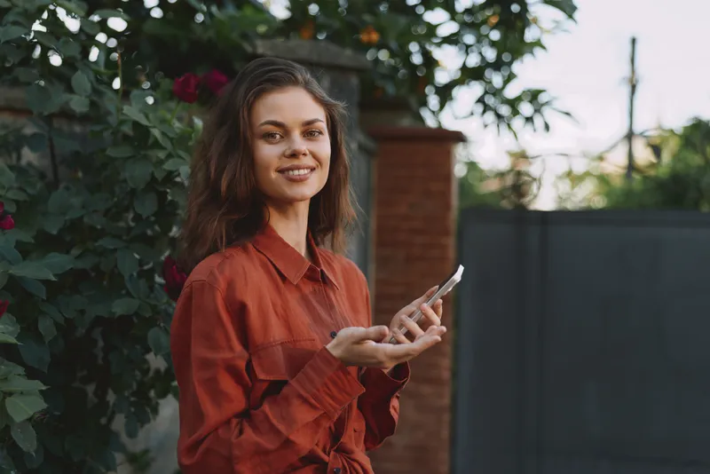 A young woman in a rust red shirt smiles while using a smartphone outdoors in a leafy residential area, conveying casual everyday connectivity and modern lifestyle