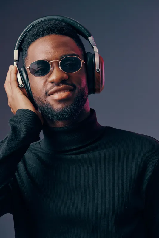 Young Black man in stylish black turtleneck and sunglasses enjoying music with headphones, expressing happiness and confidence against a gradient background