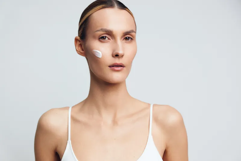 Young woman with smooth skin applying white face cream on cheek. Minimalist background and soft lighting highlight natural beauty and skincare routine concept.