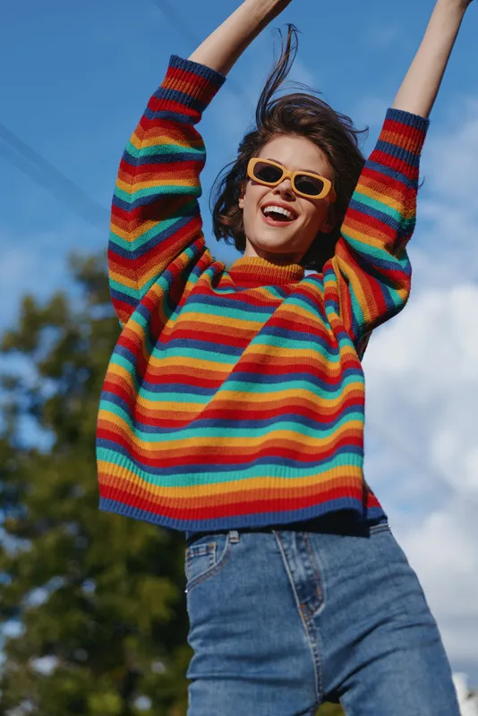 Woman sweater sunglasses smile outdoors colorful happy arms raised as young model jumps against blue sky, playful portrait in knitwear and denim with bright sunlight.