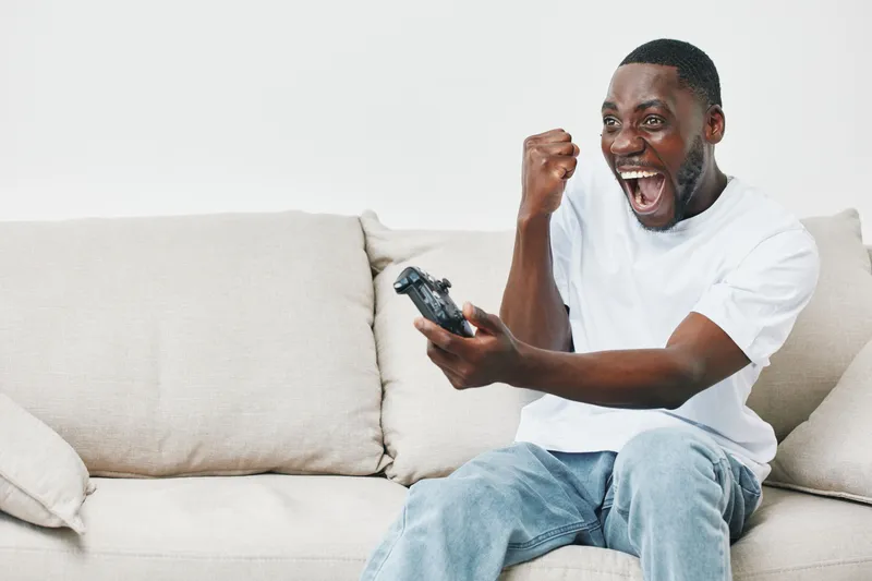 Excited young man celebrating a gaming victory at home on a cozy sofa, expressing joy and enthusiasm with a video game controller in hand