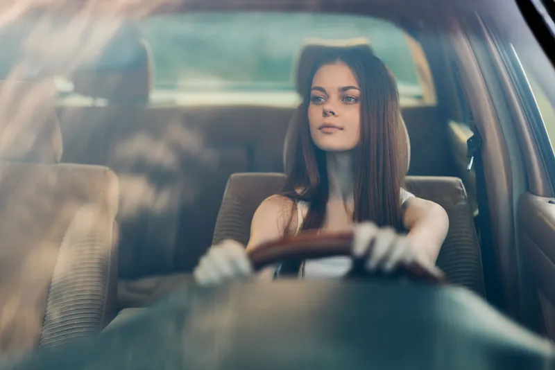 A young woman driving a car, showcasing confidence and determination, with natural light enhancing her features and a peaceful background.