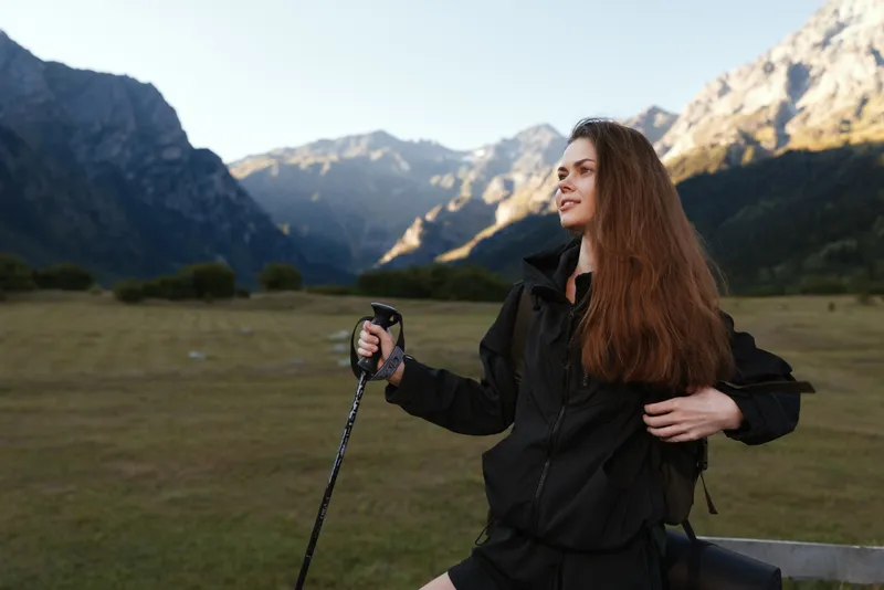 A young woman wearing a black windbreaker smiles as she holds a hiking pole in a wide valley framed by rugged mountains, conveying adventure, freedom, and outdoor exploration.