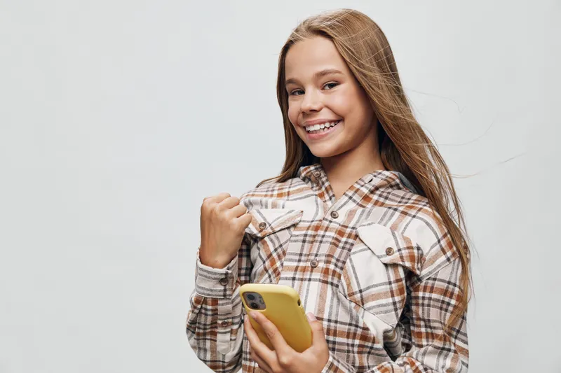 Girl smiling and holding phone in yellow case, wearing plaid shirt with long hair, standing against plain background for positive communication and modern technology.