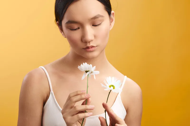 Asian woman delicately examines two white daisies in a bright studio portrait, conveying calm focus and gentle beauty against a warm background.