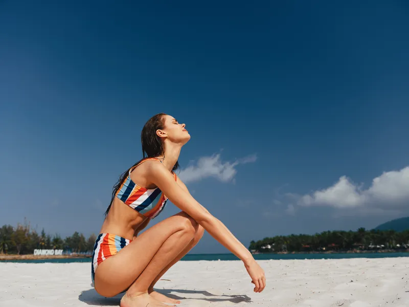 Sunlit woman crouches on bright white sand wearing a striped bikini, eyes lifted to a clear blue sky, exuding warmth, focus, and carefree seaside mood.
