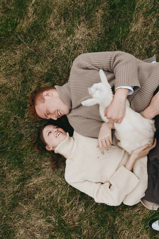 Natural, smiling couple resting on grass with a playful goat kid, celebrating outdoor lifestyle and rustic charm. Cozy sweaters, gentle light, candid affection, relaxed mood, and connection with
