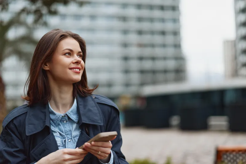 A young woman in a denim jacket stands outdoors, smiling while holding a smartphone, with a modern cityscape and glass buildings in the bright background.