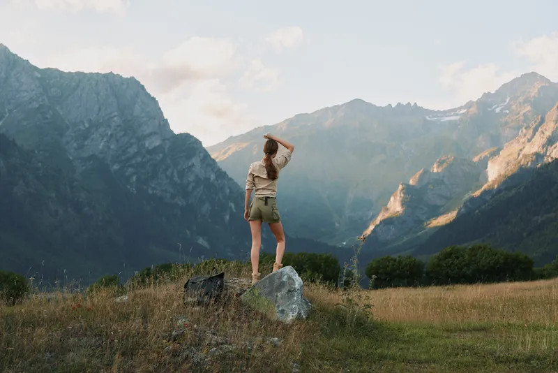 Young woman stands on a rock in a sunlit mountain meadow, gazing toward distant peaks with a sense of freedom and calm, capturing a natural outdoor moment.