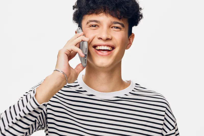 Happy man talking on smartphone wearing striped shirt, smiling with joy and excitement against white background, casual and youthful lifestyle portrait.