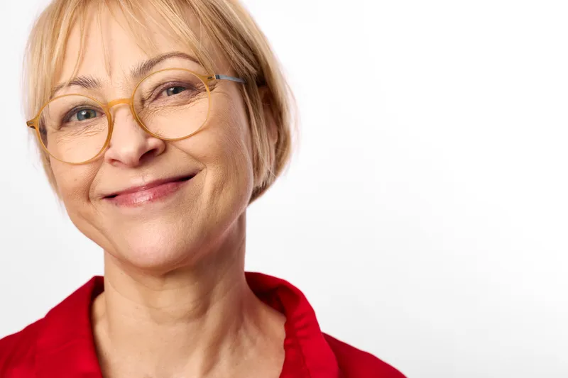 Smiling elderly woman with short blonde hair and glasses, closeup portrait, wearing red shirt, soft light, calm friendly expression, isolated on studio white background