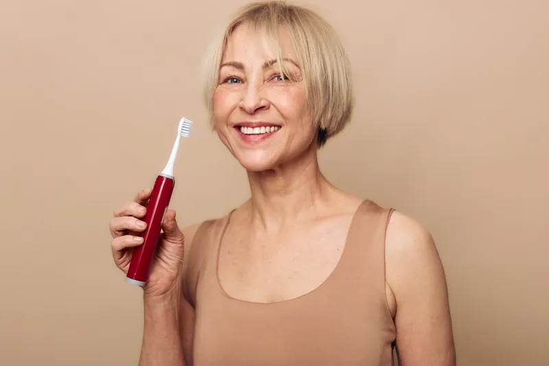 Smiling mature woman with short blonde hair holds red electric toothbrush, shows healthy teeth and confident smile on beige background. Concept of dental care and oral hygiene.