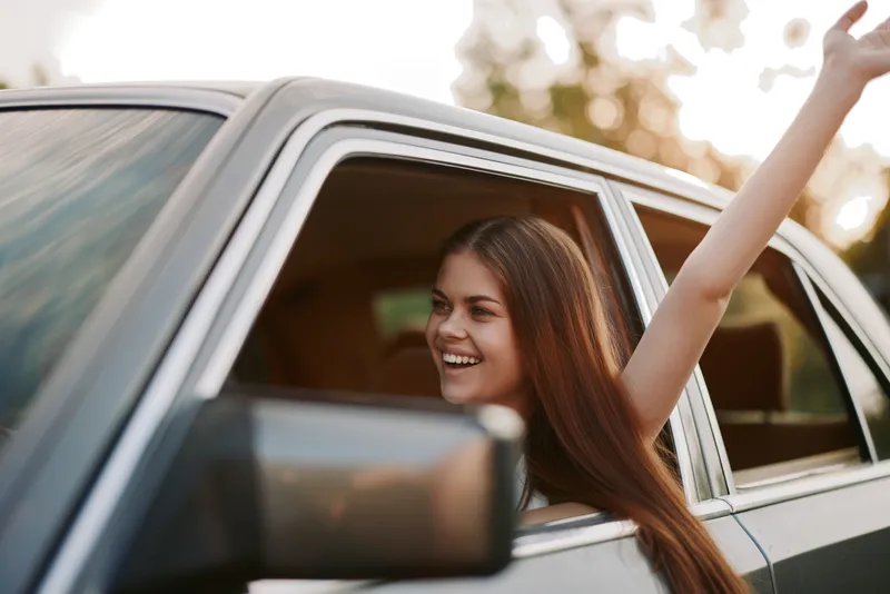 Happy young woman with long hair enjoying a car ride, raising her hand out of the window in a vibrant summer setting.