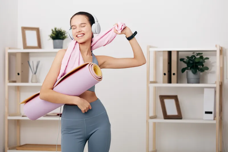 Fitness woman in a home gym, wearing headphones and a towel draped over her shoulders, holding a rolled yoga mat and smiling as she stretches after a workout.