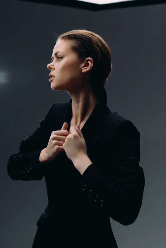 Serious woman in blazer adjusting collar with focused expression in moody studio lighting, fashion style, portrait concept