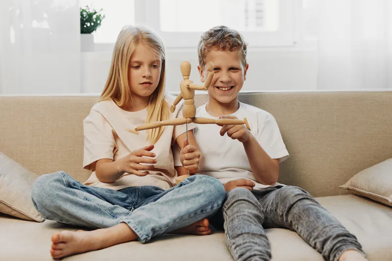 Two happy children sitting on sofa at home play with wooden mannequin toy, smiling boy and focused girl enjoy creative activity in bright living room. Concept of childhood and imagination.