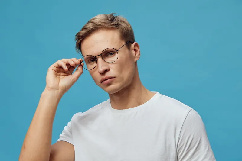Stylish young man with glasses and fair skin adjusting his glasses while looking into camera against blue background in casual white t-shirt. Portrait of intelligent, modern, poised individual for lif