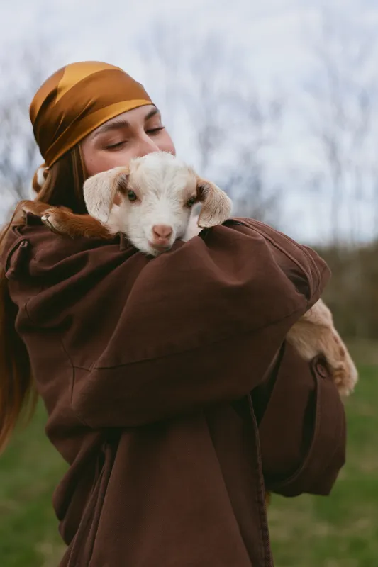 Young woman embraces a gentle goat kid outdoors, wearing a brown coat and headscarf, conveying warm rural lifestyle and tenderness, close contact with animals in a peaceful landscape