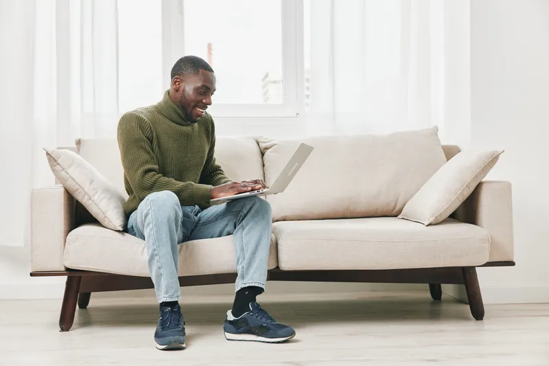 Happy African American man using a laptop on a cozy couch, showcasing modern technology and lifestyle, indoor scenery with soft colors