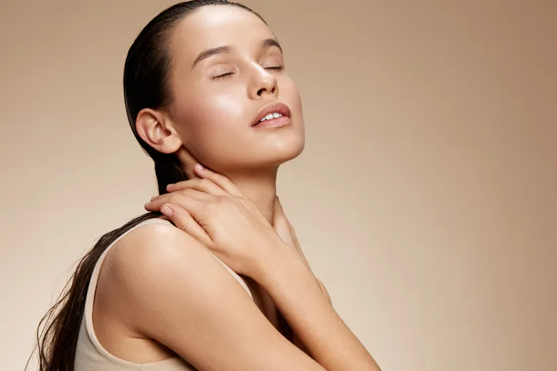 A serene young woman with smooth skin, gently holding her neck, exuding confidence and calmness against a warm beige background, showcasing natural beauty and elegance