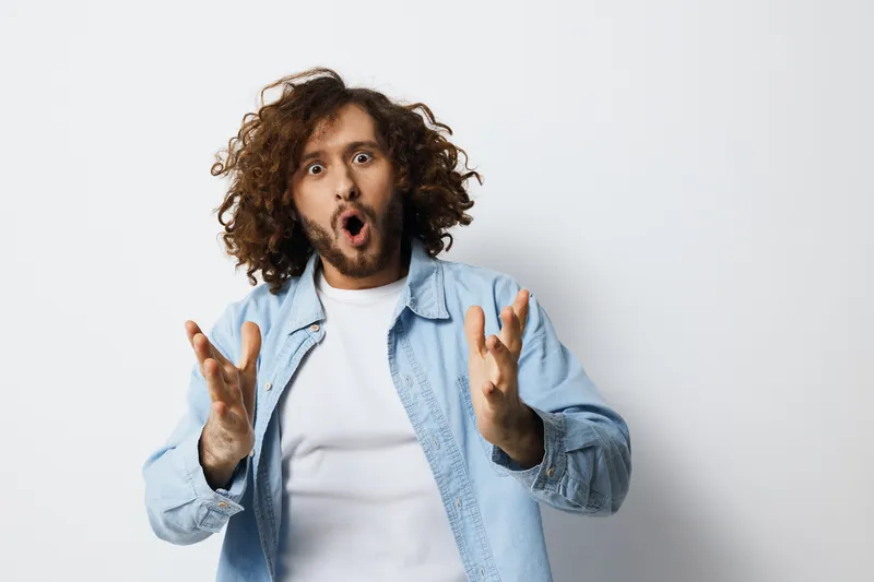 Surprised man with curly hair expressing excitement in a light blue shirt, on a clean white background, conveying a vibrant and energetic emotion for creative concepts