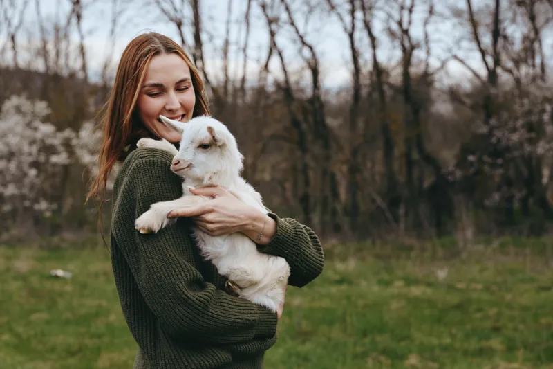 A young woman in a green sweater gently holds a white baby goat outdoors during early spring, smiling with closed eyes in a peaceful natural setting.
