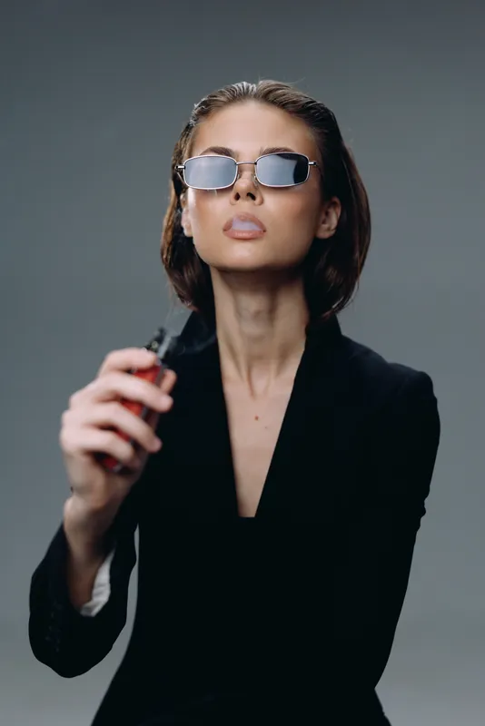Stylish woman in sunglasses holding vape device wearing black suit with confident expression