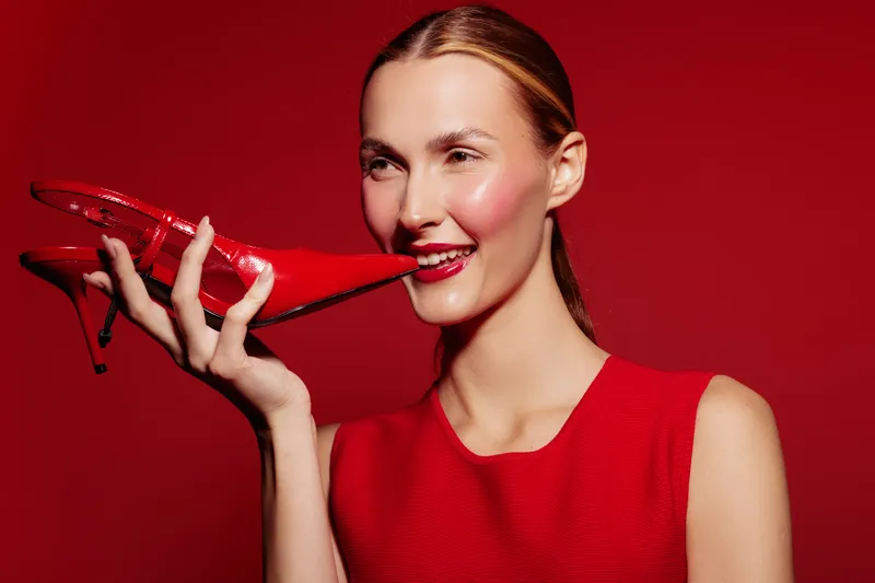 Smiling woman in red dress holds red high heel shoe near face on red background. Concept of fashion, style, beauty, and elegance with vibrant color contrast and joyful mood.