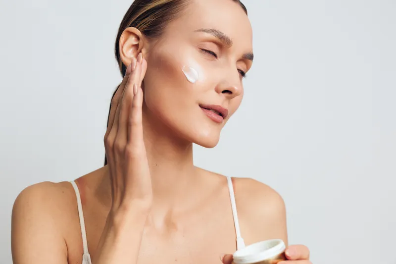 A young woman with clear skin applies cream to her face with closed eyes in a bright studio. Concept for skincare, beauty routine, and natural cosmetics care.