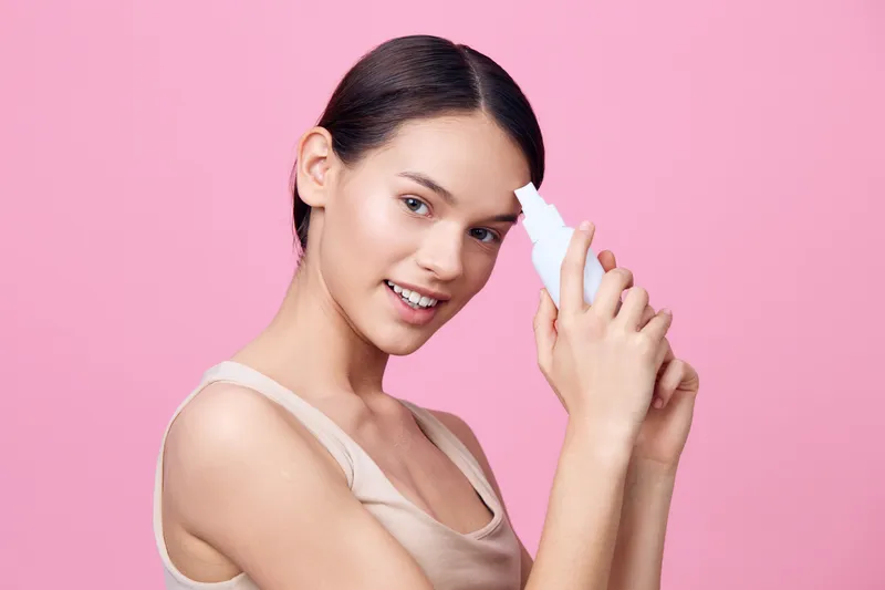 Young woman applying skincare product on her forehead, smiling against a pink background, showcasing healthy skin and beauty concept