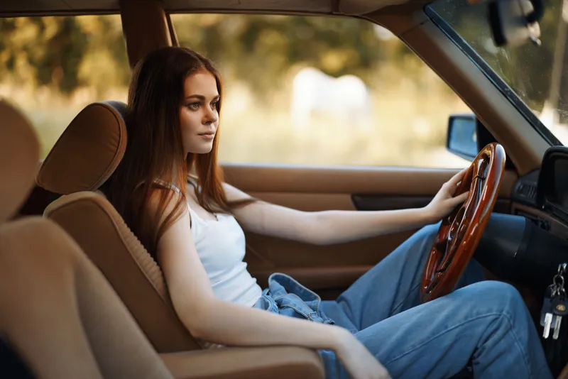 Young woman driving a vintage car with a contemplative expression, wearing a casual white top and denim jeans, surrounded by nature in a warm, autumn setting.