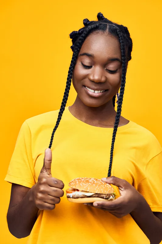 Happy young woman enjoying a delicious burger against a vibrant yellow background, showcasing a thumbs up gesture, representing a joyful eating experience