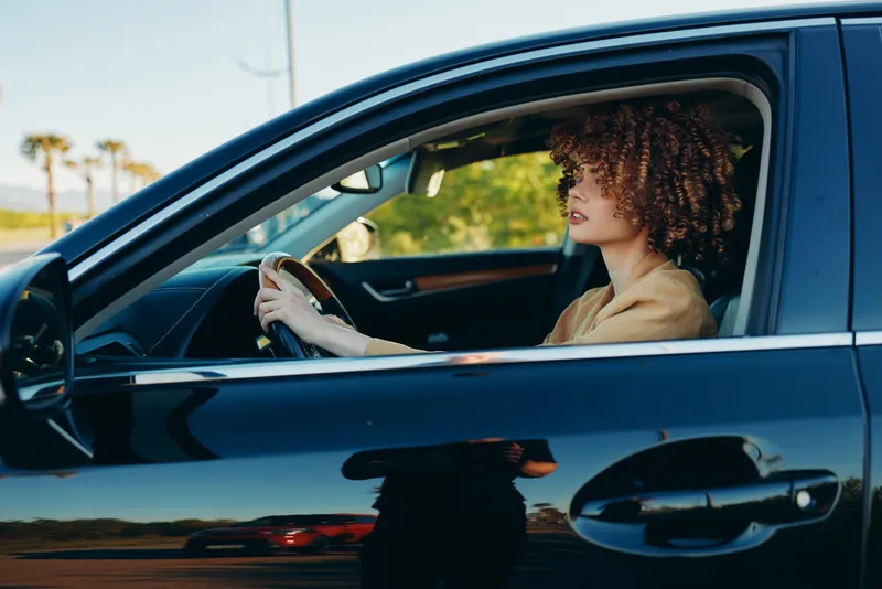 Curly haired woman driving a car, hands on the wheel, calm expression in a lifestyle portrait. Natural light and warm retro film color create a relaxed, everyday journey mood.