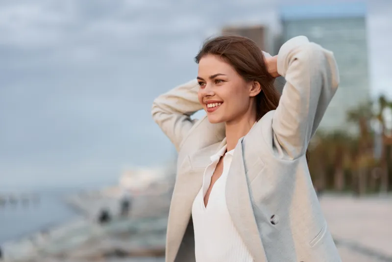 Young woman in beige blazer enjoying city waterfront with smile, casual fashion style, outdoor lifestyle, relaxed confident mood, urban background