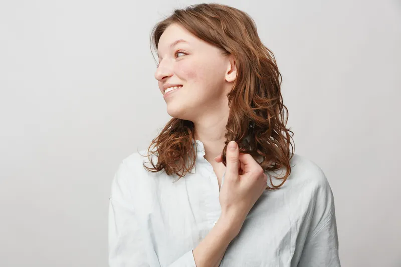 Young woman with curly hair smiles gently looking sideways, wearing a white shirt. Studio portrait on light background with natural expression and casual style.