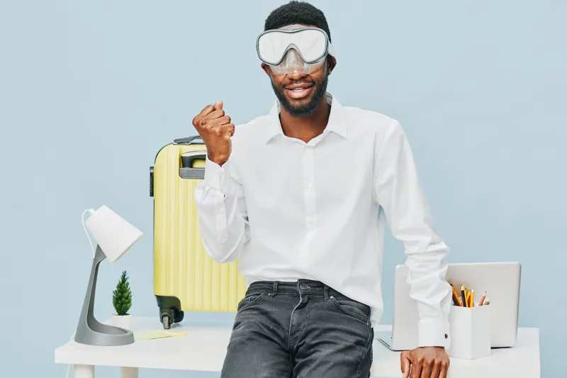 Smiling man with beard wearing protective goggles, white shirt, and dark jeans, celebrating success while standing in modern workspace with travel luggage, desk lamp, and pens, bright background, life