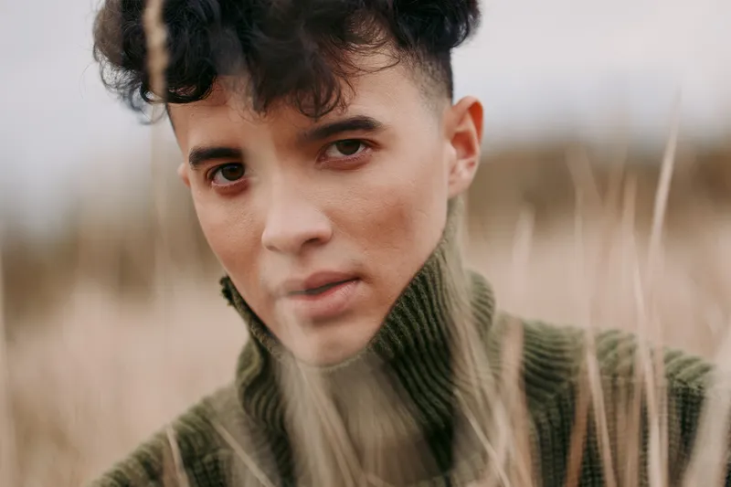 Close portrait of a young person in a grassy field, wearing a knit turtleneck, soft natural light revealing an authentic, unposed expression and genuine, quiet presence.