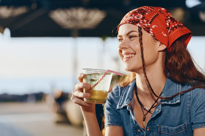smiling woman with red bandana enjoying a cold drink outdoors in warm sunny weather, wearing denim jacket and casual summer style