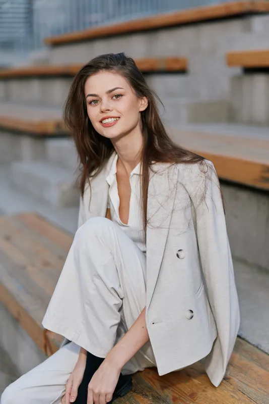 Confident businesswoman smiling in a light-colored blazer and pants, exuding professionalism and charm in an urban setting with a warm color palette.