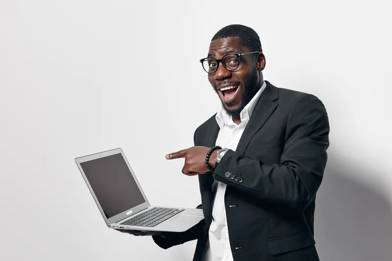 African American man with dark skin wearing glasses and black suit holding laptop and pointing at screen with excitement on white background in studio.