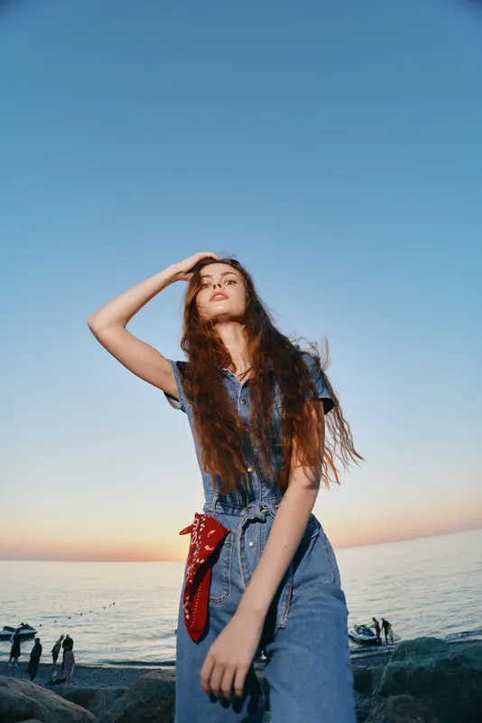 Lifestyle woman in modern boho-western denim street style posing by the sea at sunset, film color effect with natural light and relaxed mood.