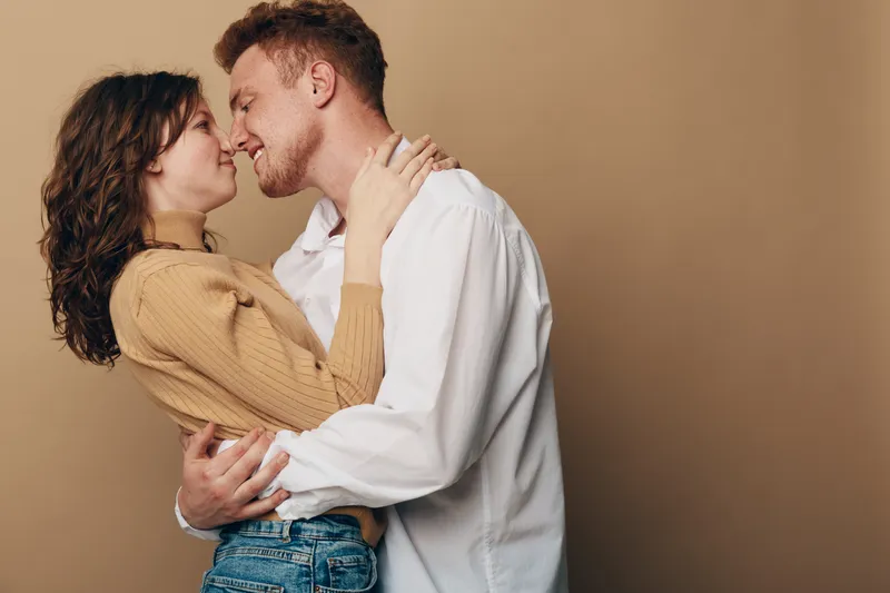 Young couple in casual clothes embraces and leans in for a kiss against beige background. Intimate moment, love, affection, and romantic connection between man and woman.
