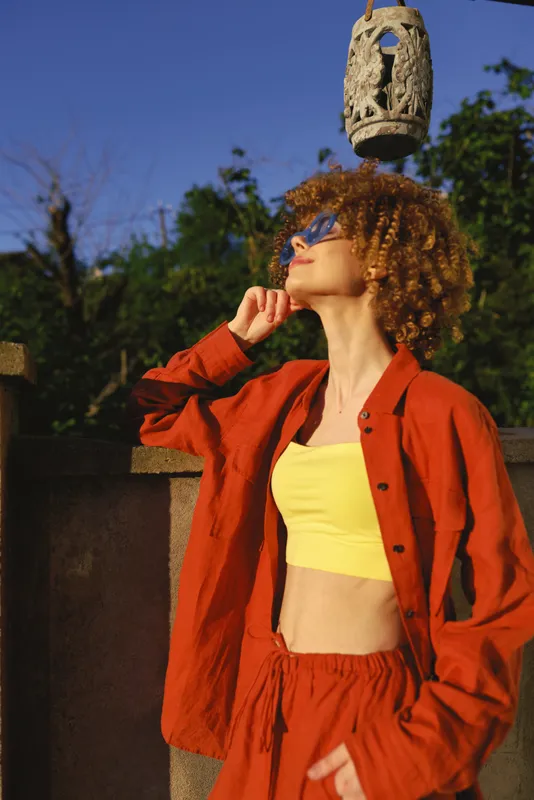 Woman with curly hair wearing an orange jacket and yellow top stands outdoors under a blue sky with a hanging lantern, conveying a fashionable, relaxed lifestyle and sunny mood