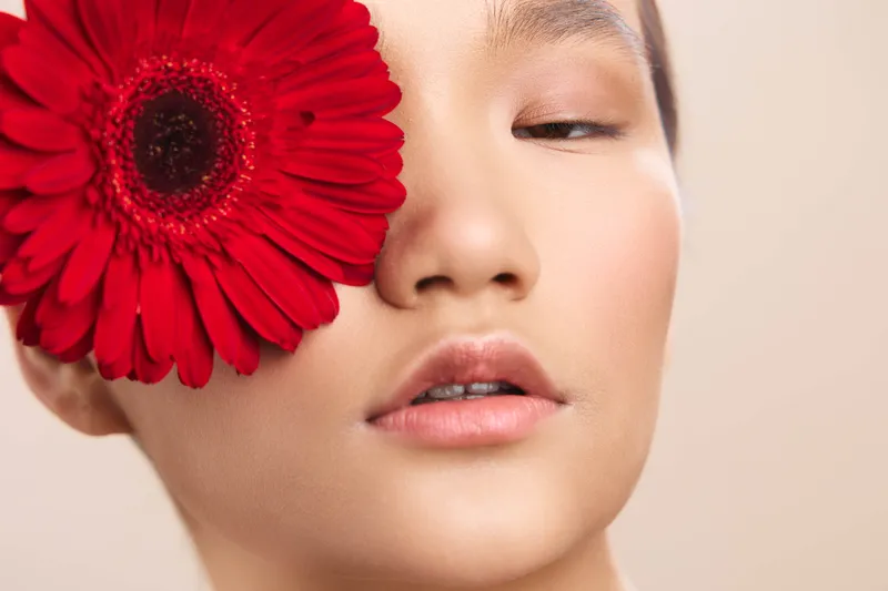 asian woman with a vibrant red flower held near her eye, soft lighting highlights flawless skin, creating an artistic portrait that conveys elegance, serenity, and quiet confidence.