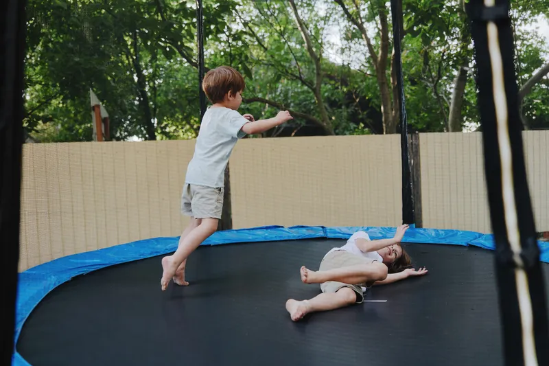 Energetic boy jumping on a trampoline, playing joyfully outdoors with a friend, surrounded by green trees, capturing the essence of childhood fun and freedom.